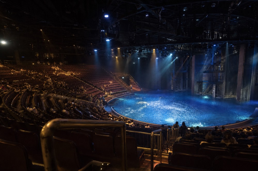 Tiered seating rows curving around a lit water stage seen from the upper bowl