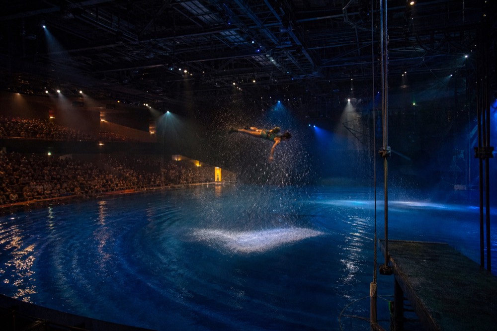 Performers mid-dive above a large stage pool lit in blue