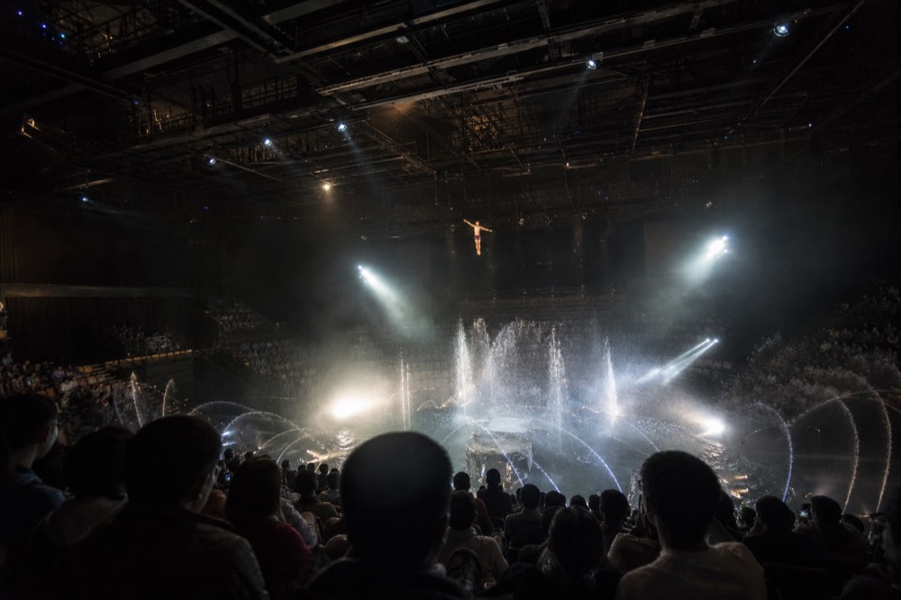 A high diver mid-air above the water stage with fountain spray rising around the platform